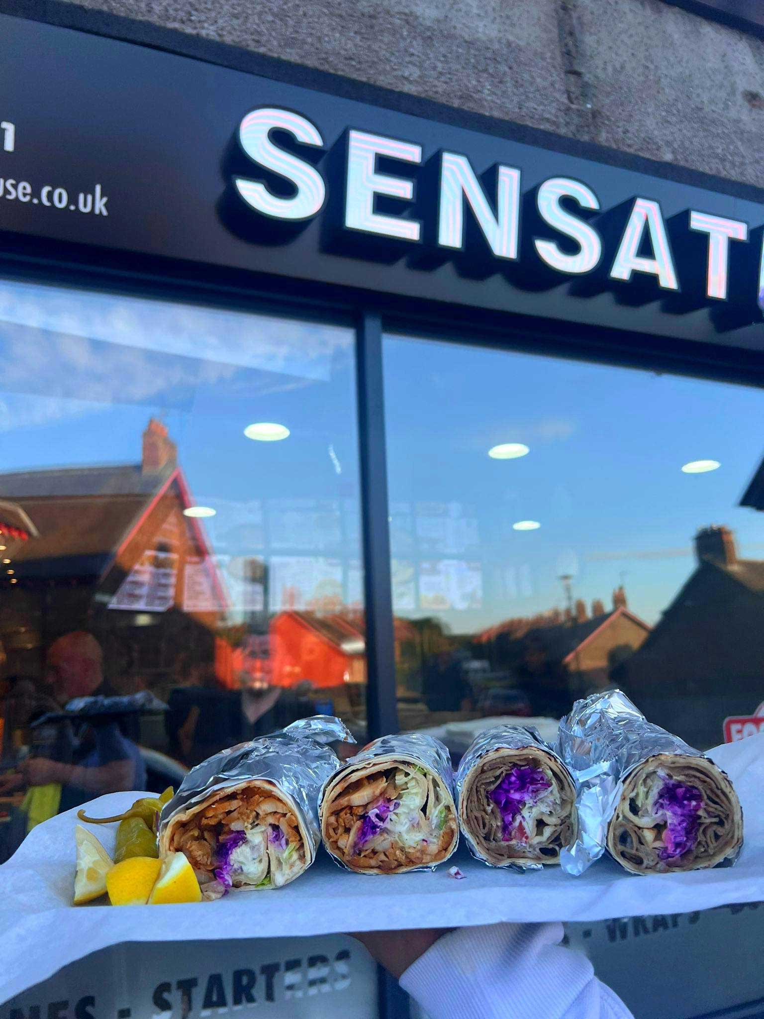A tray with four foil-wrapped kebabs is held in front of Sensation Takeaway.
