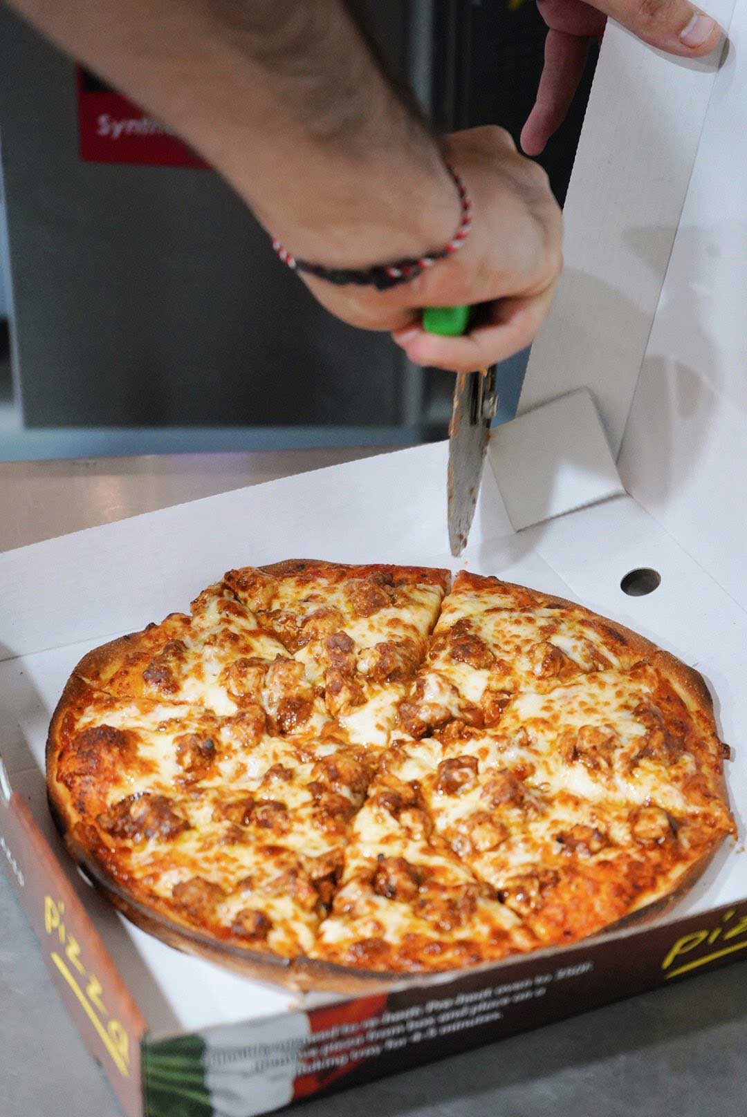 A knife slices a chicken and cheese pizza inside a Sensation takeaway’s cardboard pizza box.